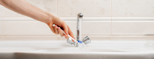 a close up hand holding a white toothbrush and wash it under the water in sink in bathroom