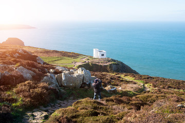 A view of the Welsh coastline, with blue sky and bright, hazy sunshine. There is a white building and a man down below, taking a photo of the view