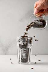 Iced coffee concept. A male hand pours coffee beans into a tall glass with ice. Copy space.
