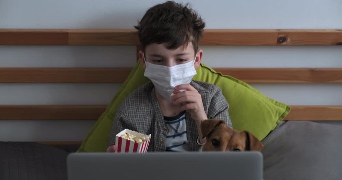 Boy medical mask face during quarantine watches movie enthusiasm, throws popcorn in mouth out box, misses target, dog Jack Russell Terrier. Coronavirus COVID-19 virus. Epidemic, pneumonia, pandemic