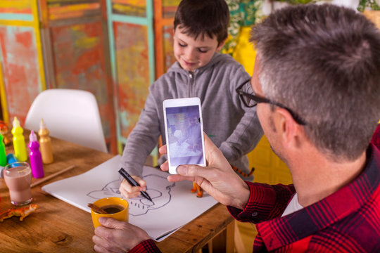 Father taking picture of his son while the child draws.