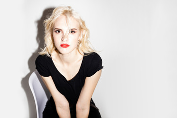 Fototapeta premium young beautiful blonde girl in a black t-shirt and black jeans sitting on a chair and looking at the camera on a white background, studio fashion photo with copy space