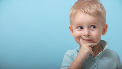 a three-year-old kid in a blue Polo poses for the camera and shows different emotions. emotions of children. children on a blue background. the boy thinks and holds a finger to his face