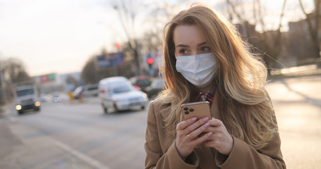 Caucasian young woman in medical mask texting message and tapping on mobile phone at street with road and cars. Girl typing and scrolling on smartphone in city outdoors. Pandemic healthcare concept.