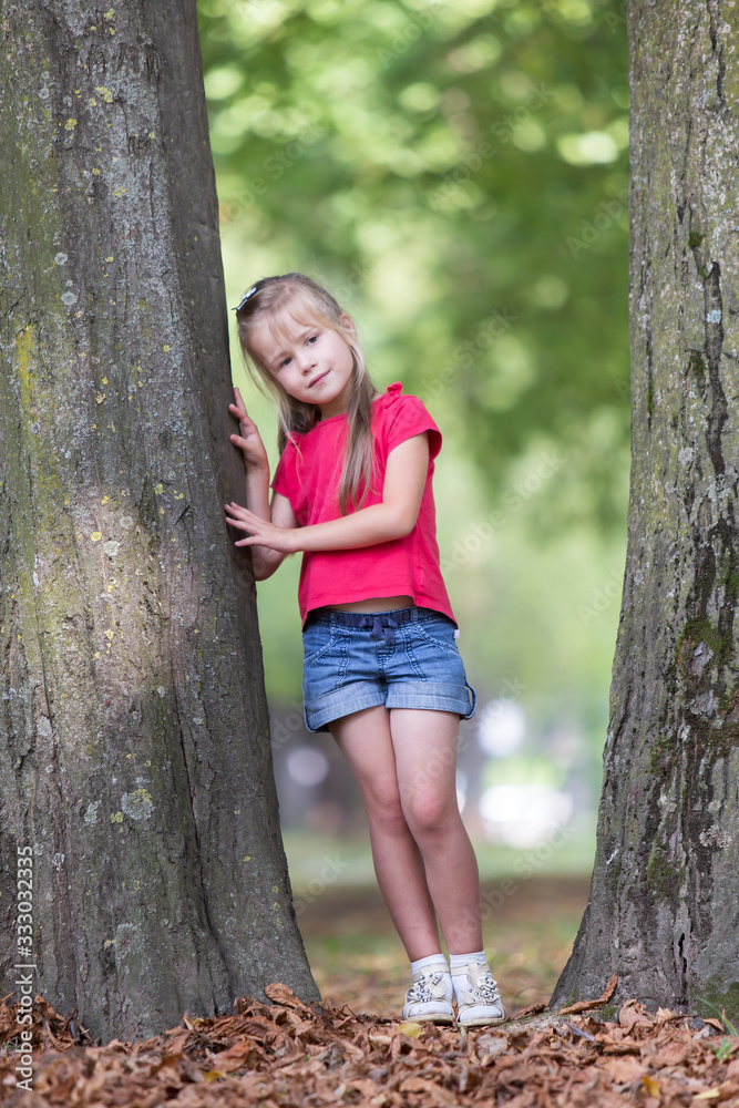 Wall mural Portrait of a pretty little child girl standing near big tree trunk in summer park outdoors.