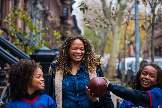 Happy girl holding American football while standing by sister and mother on footpath in city