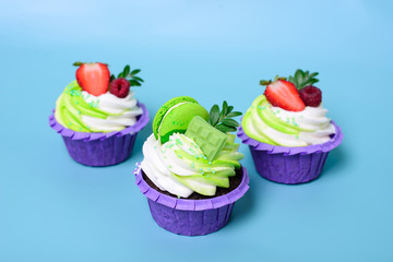 Three cupcakes with whipped cream, chocolate bar, strawberry ,decorated macaroons on blue background. Picture for a menu or a confectionery catalog. with space for text.