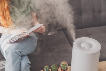 Household humidifier at home on table near woman reading on sofa - obrazy, fototapety, plakaty