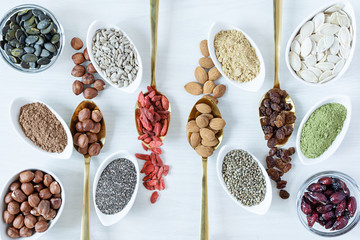 Golden spoons and white bowls with various superfoods on white wooden background top view.