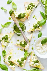 Crispy toasts (crostini, bruschetta) with cottage cheese (ricotta) and freshly prepared pesto. Homemade basil pechto sauce in a jar on a light background.