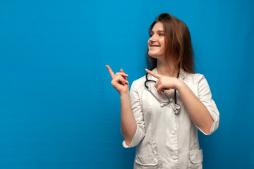 young cheerful girl nurse smiles and points a finger at a place for text, female doctor on a blue background