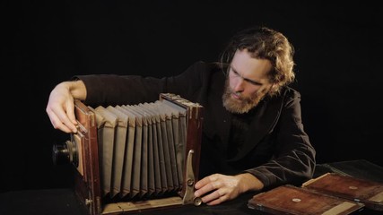 Image of curious european man of Dostoevsky in black coat, using and checking old 19th-century camera on dark background. Bearded Russian photographer works with antique camera in black isolated room.