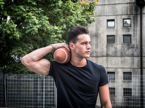 Handsome young man standing on street and holding American football ball.