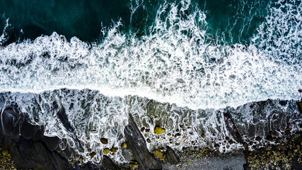 Aerial view on crashing waves with colourful stones on black sand beach. Abstract background. Green dark waters.