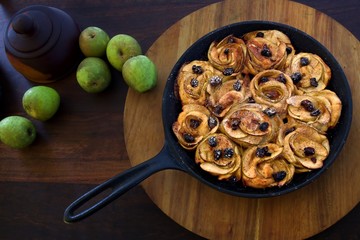 Baked apple rosettes in pan on a wooden board on a wooden table.  Green apples are scattered to the side across the table and a wooden pot sits aside the apples