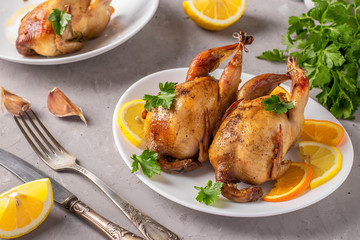 Baked quails with lemon and orange served on a white plate on a gray background