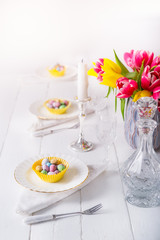 Serving Easter table with colorful sweet eggs in cases on the plates, crystal glassware, and fresh tulip flowers on the white wooden table. Family holiday background. Light vertical card. Copy space.