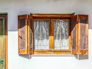 Old wooden window with open shutters on white stucco wall with insect netting tacked on and white lace peacock curtains - part of door visible - golden hour sun and shade
