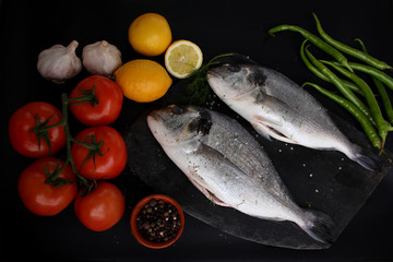 lemons, tomatoes, hot peppers and two carcasses fresh raw atlantic saltwater fish Sparus aurata with shiny silver scales lie on a black background, cooking concept