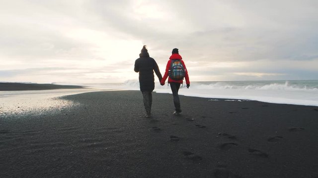 Young hipster couple walking on volcanic black sand beach in Iceland and having fun at sunset, slow motion