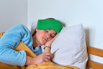 Sick girl with forehead compress is laying in bed under blanket and holding a thermometer to check...