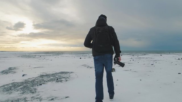 Young man traveler with camera runs to the ocean on snow desert in Iceland. Slow motion shot at sunset or sunrise.