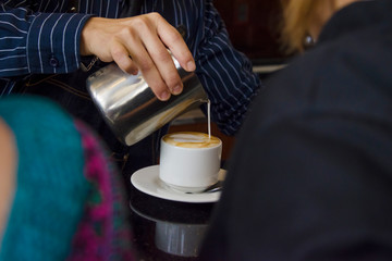 Fototapeta na wymiar Mano de barista dibujando una flor sobre la espuma de café mientras mujeres esperan