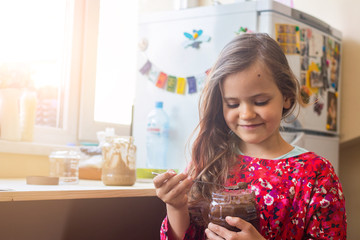 Cute happy young smiling girl eat homemade sweet chocolate paste spread from big glass jar with joy...
