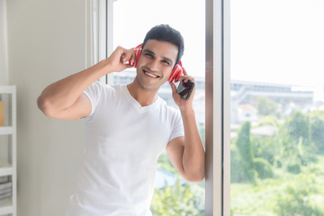 An Indian handsome man is listening to music from his headphones connected wireless to his mobile phone. He has a smiling face and looks happy in the living room in the morning.