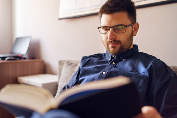 One adult caucasian man sitting on the sofa bed at home reading the book study learning alone during the quarantine lockdown in a day wearing shirt and eyeglasses and beard front view