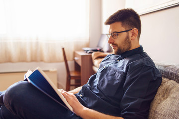 Naklejka premium One adult caucasian man sitting on the sofa bed at home reading the book study learning alone during the quarantine lockdown in a day wearing shirt and eyeglasses and beard side view