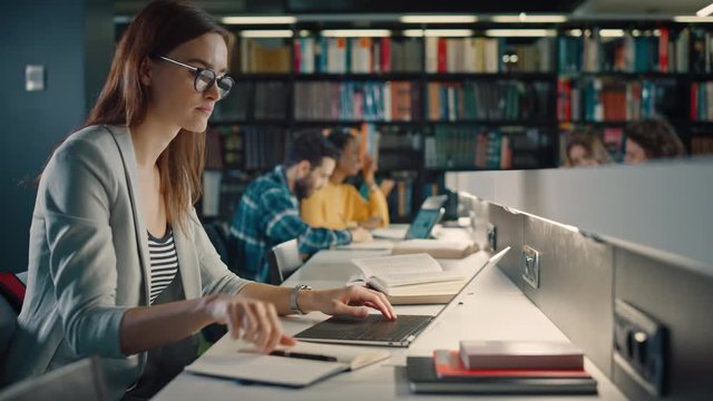 University Library: Talented Caucasian Girl uses Laptop, Smart Helpful Classmate Explains and Advices Her with Class Assignment. Happy Diverse Students Talking, Learning, Studying Together for Exams