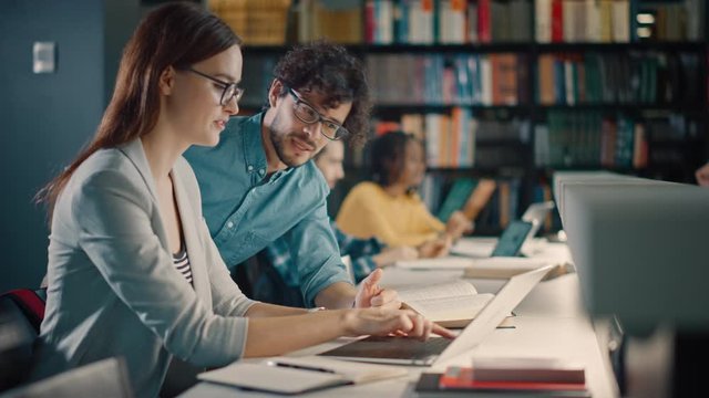 University Library: Talented Caucasian Girl uses Laptop, Smart Helpful Classmate Explains and Advices Her with Class Assignment. Happy Diverse Students Talking, Learning, Studying Together for Exams