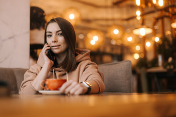 Young woman freelancer in casual clothes talking on the phone at a table in a cozy cafe. Coffee cup on the table. Concept of leisure activity.