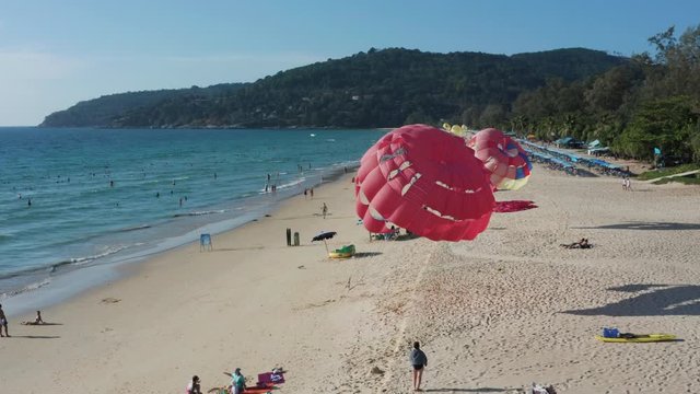 Tropical beach with numerous tourists having sun tan and swimming in water, with several parasails installed on shore on a sunny day. Karon Beach, Phuket, Thailand.