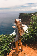 Beautiful girl traveler with long hair and a slender body stands on a cliff of a cliff overlooking the waves of the blue ocean.Bali Island, Indonesia. Travel and tourism concept in exotic and tropical