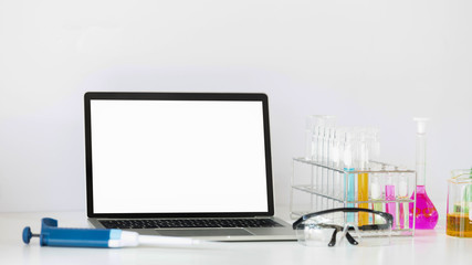 Photo of Scientific experiments equipment putting on white working desk with white blank screen computer laptop. Flat lay computer laptop, chemistry glassware, safety glasses.