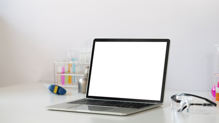 Photo of Scientific experiments equipment putting on white working desk with white blank screen computer laptop. Flat lay computer laptop, chemistry glassware, safety glasses.
