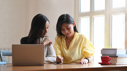 Young women talking together while sitting at the wooden working table with computer laptop and tablet next to living room windows as background.