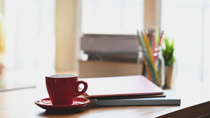 Coffee morning concept, Red coffee cup with coasters putting on wooden table with pencil holder, stack of books, potted plant over living room windows as background.