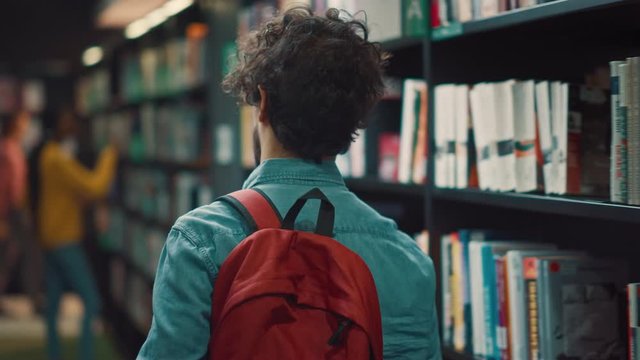 University Library: Student Walks Between Rows of Bookshelves Searching for the Right Book Title for Class Assignment and Exam Preparations. Back View Following Camera Shot. Young People Study, Learn