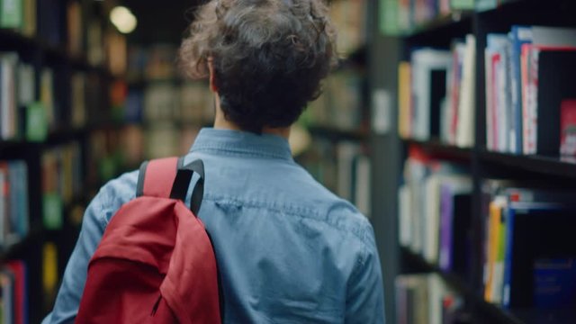 University Library: Student Walks Between Rows of Bookshelves Searching for Right Book Title for Class Assignment and Exam Preparations. Back View Following Slow Motion Camera Shot. Young People Study
