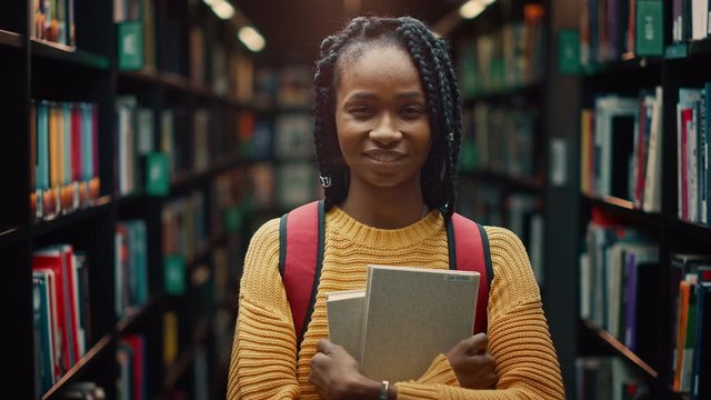 University Library: Smart Beautiful Black Girl Standing Next to Bookshelf Holding and Reading Text Book, Doing Research for Her Class Assignment, Exam Preparations. Authentic Students Study, Succeed
