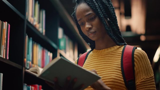 University Library: Smart Beautiful Black Girl Standing Next to Bookshelf Holding and Reading Text Book, Doing Research for Her Class Assignment and Exam Preparations. Low Angle Portrait with a Smile