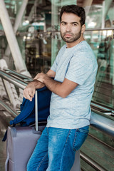 Fototapeta na wymiar Thoughtful young man leaning on metal railing at airport. Serious young man with luggage at terminal. Travel concept