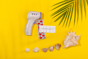 Top view on infrared thermometer, white and red pills in blister pack, seashells, palm leaf on...