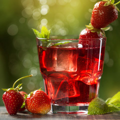 red strawberry drink in a glass, with the addition of mint and ice, next to it are strawberries and mint leaves, against a background of greenery