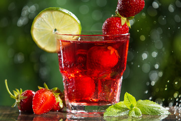 refreshing red strawberry drink in a glass, with the addition of lime, mint and ice cubes, next to it are strawberries and mint leaves, against a background of greenery, drops of water and splashes