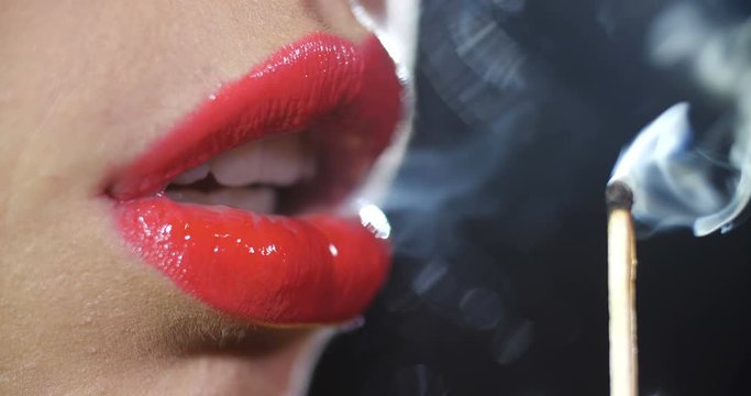Detail close-up macro shot of the lips of a girl painted with a shiny red lipstick  blowing out the fire of a burning match on black studio background isolated. 4k slow motion 50 fps