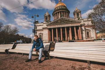 A young blond man in a jacket sits on a white bench with a panoramic view of St. Isaac's Cathedral